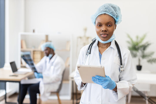 Female African doctor wearing protective medical gear standing in clinic holding tablet device. Background nurse sitting at desk in white coat working.