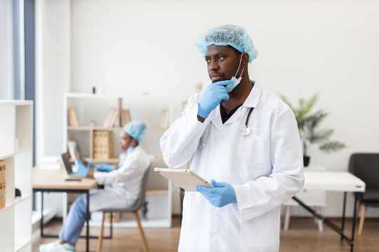 Male adult healthcare professional in medical office wearing scrubs, blond hairnet, gloves and stethoscope, holding clipboard, contemplating work with background colleague on laptop.