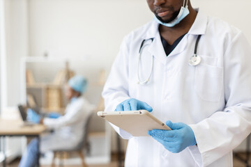 Male adult doctor wearing white coat and gloves, working on digital tablet, medical setting with healthcare professional in blurred background, focused on patient care technology