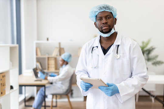 Confident African male doctor in surgical attire holding tablet in modern hospital environment. Female medical staff member working in background providing professional healthcare