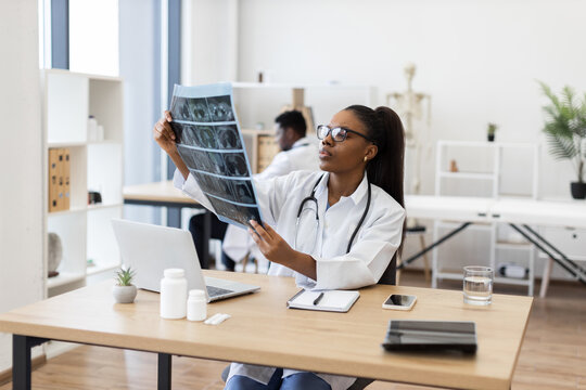 Confident young adult female doctor evaluating X-ray scans in office workspace. Medical professional focusing on diagnostics and patient records. Modern healthcare settings emphasizing professionalism