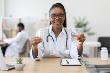 Young Black female doctor holding blister pack with medicine and glass of water. Sitting at desk in medical office, wearing white coat with stethoscope.