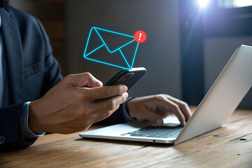 Close up of a businessman checking his smartphone for new email notifications while working on his laptop at a wooden desk with a glowing envelope icon
