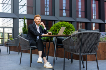 Smiling red-haired businessman in suit working on laptop at outdoor cafe