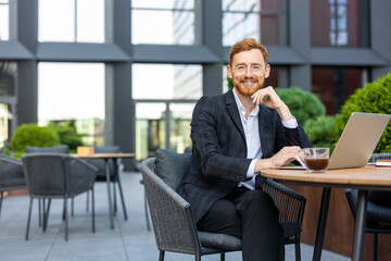 Smiling businessman with ginger hair sitting at urban cafe with laptop