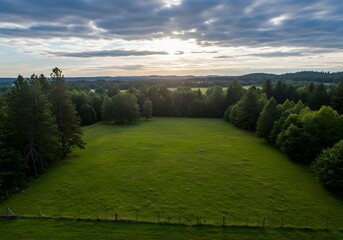 Obraz premium Aerial view of lush green meadow surrounded by trees under a cloudy sunset sky.