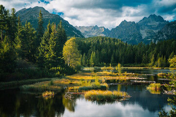 Fototapeta premium As the sun sinks behind the towering peaks, the quiet lake reflects the sky in shades of orange and gold, offering a moment of untouched silence in the heart of the mountains.