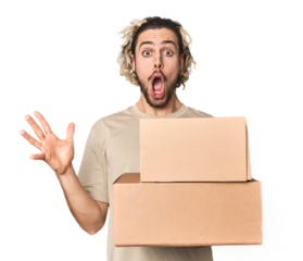 Young man with cardboard boxes in studio receiving a pleasant surprise, excited and raising hands.