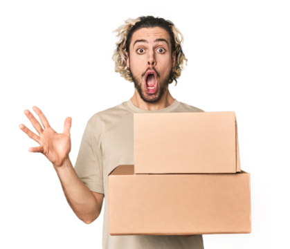 Young man with cardboard boxes in studio receiving a pleasant surprise, excited and raising hands.