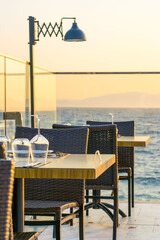 Elegant restaurant terrace with neatly arranged tables and chairs overlooking the sea at golden hour. Luxury seaside dining experience with glass railing, mountain silhouettes in the background, and