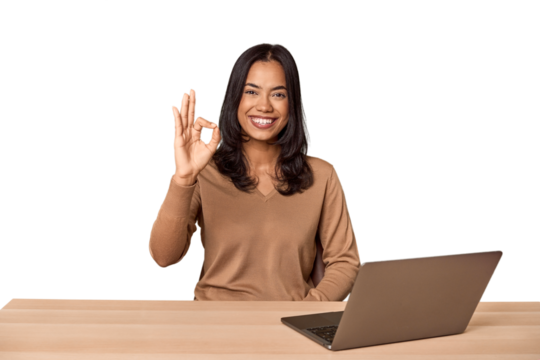 Filipino woman at desk with laptop cheerful and confident showing ok gesture.