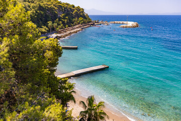 Obraz premium The Aegean coast on a sunny summer day in Kusadasi. Azure water, piers, green hills, palm trees and a promenade with the flag of Turkey. A picturesque seascape, a popular tourist destination for
