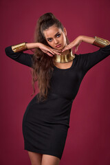 Beautiful brunette with luxurious long hair in black cocktail dress and Greek gold jewelry posing in studio on red background