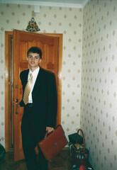 Young man in suit holding briefcase on first day of university, analog photo taken in home hallway early 2000s Ukraine with vintage atmosphere