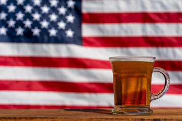 Glass of beer on wooden table against the backdrop of an American flag, closeup. The most popular...
