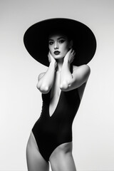 Beautiful fashion model in swimsuit and hat posing on studio background, black and white