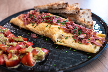Healthy breakfast with folded omelette, fresh tomato salad with avocado dressing, and whole grain toast served on black plate, balanced meal rich in protein and fiber.