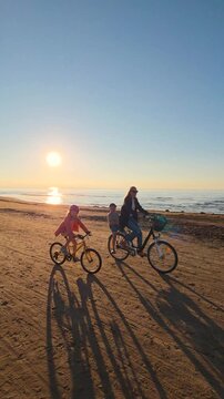 family with children riding with bicycles on beach sand at sunset