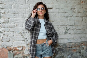 A fashionable woman poses casually, wearing sunglasses and a plaid shirt against a brick wall.
