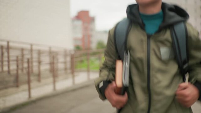 close up of student running holding notepad under arm with backpack on shoulder blurred view of college building iron frame and greenery in background showing energetic academic lifestyle