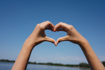 Hands forming heart shape against blue sky and lake in summer