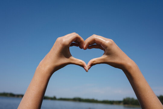Hands forming heart shape against blue sky and lake in summer