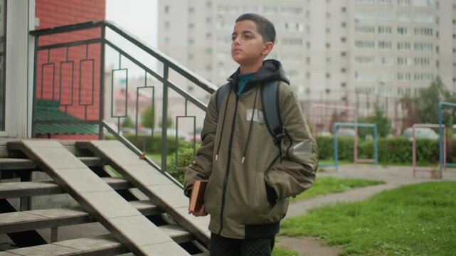 Calm kid with backpack carrying book walks through outdoor area toward bench preparing to sit and read with focused expression surrounded by grass, playground frames and urban buildings