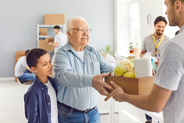 Fototapeta premium Sad elderly man and child receive a box of food from a volunteer, close-up. Group of volunteers working in a warehouse. Concept donation bag, charity foundation, community service