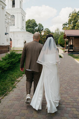 Groom in classic suit holding hand of bride in white dress as they walk near church on their...