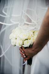 Bride in a white dress holds a bouquet of white roses in her hands behind her back covered with a...