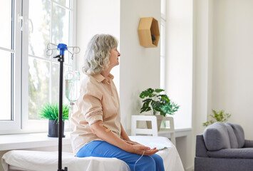 Senior woman patient receiving intravenous vitamin therapy at the clinic. Gray haired old lady with a catheter needle in her vein sitting on a hospital bed. Health care, treatment concept