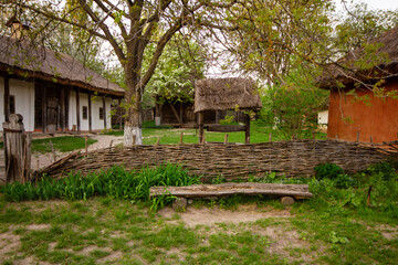 A traditional rural house and well in a rustic village setting, representing historical architecture and countryside life. The image features a wooden fence, a simple wooden bench, and traditional tha