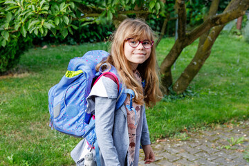 Happy little elementary school girl with eyeglasses and satchel backpack walking outdoors on the...