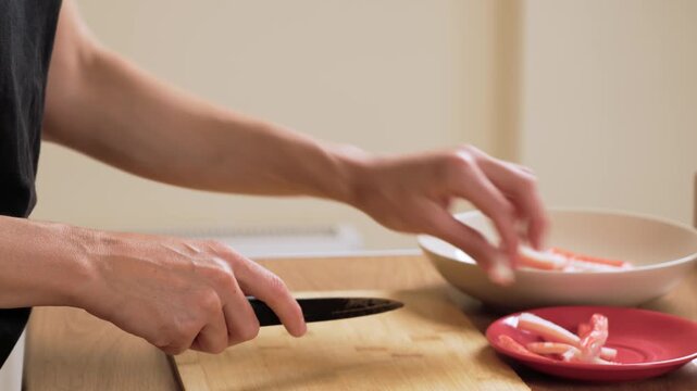 Cutting crab stick on wooden board with knife. Unrecognizable person slicing imitation seafood into pieces. Preparing crab stick portions for cooking process. Getting crab stick filling ready for