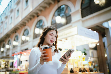A stylish young woman, possibly an influencer, holds a coffee cup and uses her smartphone outdoors in an urban cafe environment.