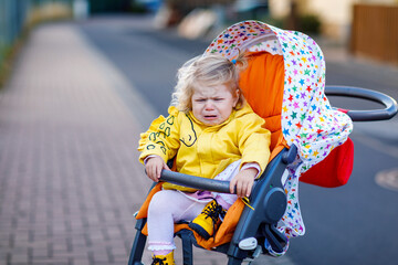 Portrait of little sad toddler girl sitting in stroller and going for a walk. Crying baby child...