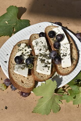 Close-up bread slices with cottage cheese and grapes on plate on parchment paper among grapes and leaves, top view