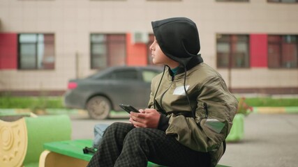 Teen boy in hoodie jacket sits on green bench holding smartphone, looking to the side with thoughtful expression while car passes by in blurred urban street background - Powered by Adobe