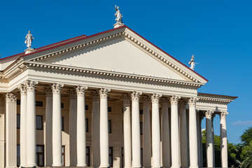 Neoclassical winter theatre building features impressive portico with tall Corinthian columns, pediment decorated with statues and red tile roof against clear blue sky. Sochi, Russia - June 28, 2025