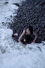 Young woman in a sparkly black dress by the ocean shore, exploring the rocky beach.