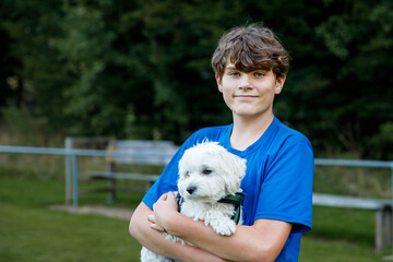 Happy teenager boy holding cute Maltese puppy dog on arms, outdoors. Smiling young male enjoying...