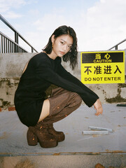 Stylish woman in a black dress and shiny boots poses near caution sign outdoors Chinese word means...
