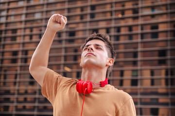 Confident young man standing outdoors in front of a modern building, raising his fist in the air as...
