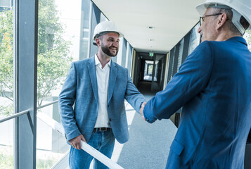 Architects shaking hands in modern office corridor wearing safety helmets
