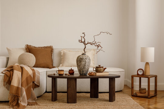 Cozy living room with beige sofa, neutral pillows, plaid blanket, ceramic vase with branches and rustic coffee table decor.
