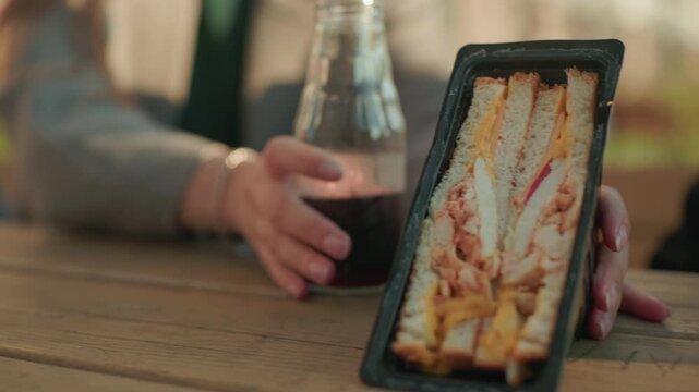 Close up of sandwich pack filled with meat and cheese placed on wooden table beside glass bottle of juice while hands move it, warm outdoor setting with natural daylight atmosphere