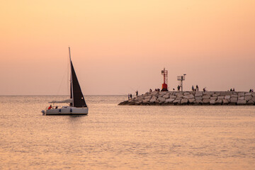 Yacht ship is sailing. Lighthouse in Rimini and fishermen on the pier. Calm on the coast. The sky...
