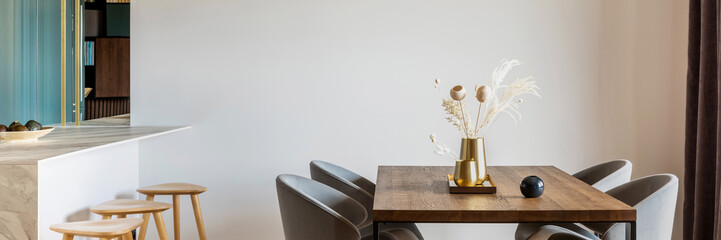 Modern and vintage dining room with brown wooden table, grey chairs and stylish chandelier. Minimalistic white wall. Creative accessories.