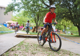 Naklejka premium Focused boy in red shirt riding bicycle on park track with friends, enjoying outdoor sports, competition and childhood adventure