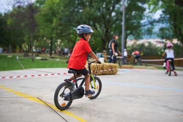 Smiling young boy riding bicycle with helmet on park track surrounded by children, enjoying outdoor...